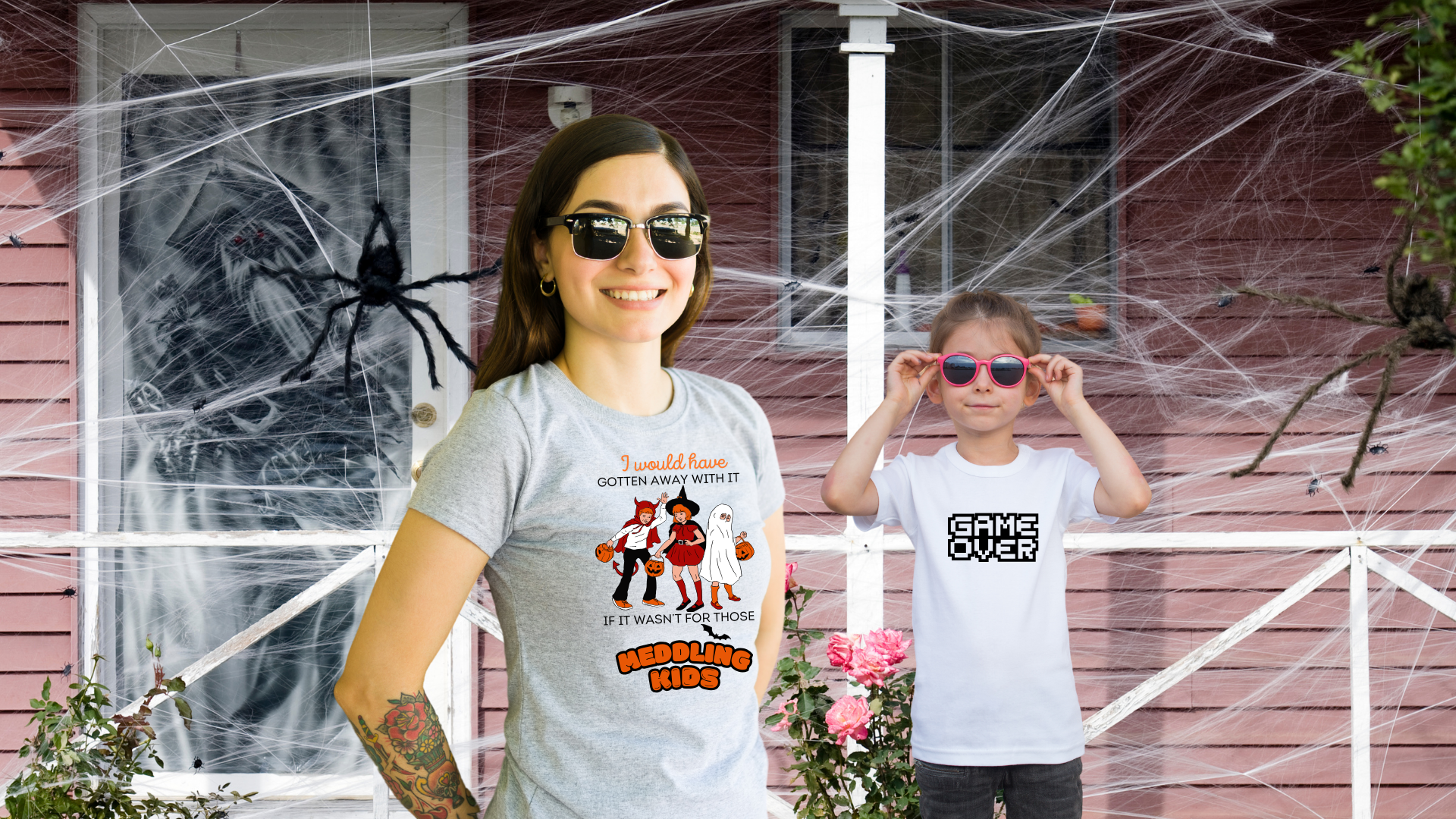 Woman wearing a 'Meddling Kids' Halloween-themed t-shirt and a child wearing a 'Game Over' t-shirt, standing in front of a spooky, decorated house.